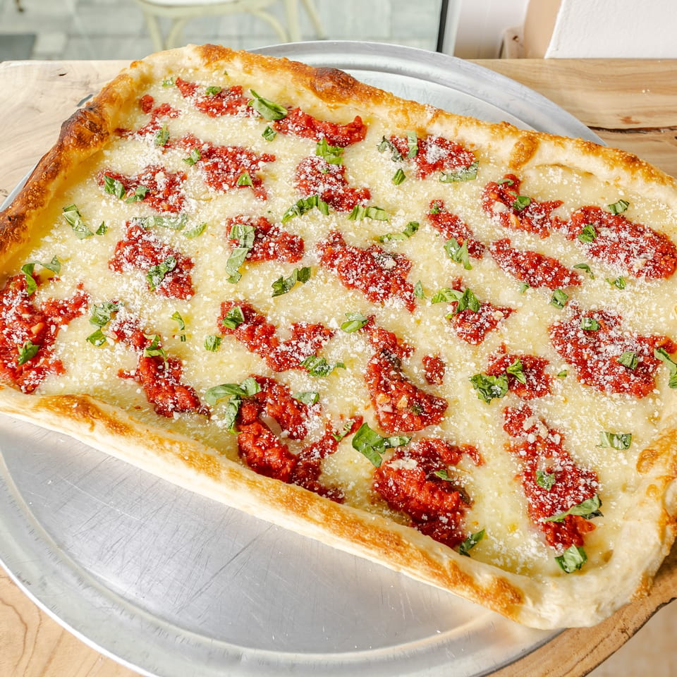 Large rectangular pizza topped with melted cheese, tomato pieces, grated Parmesan, and fresh basil on a metal tray.