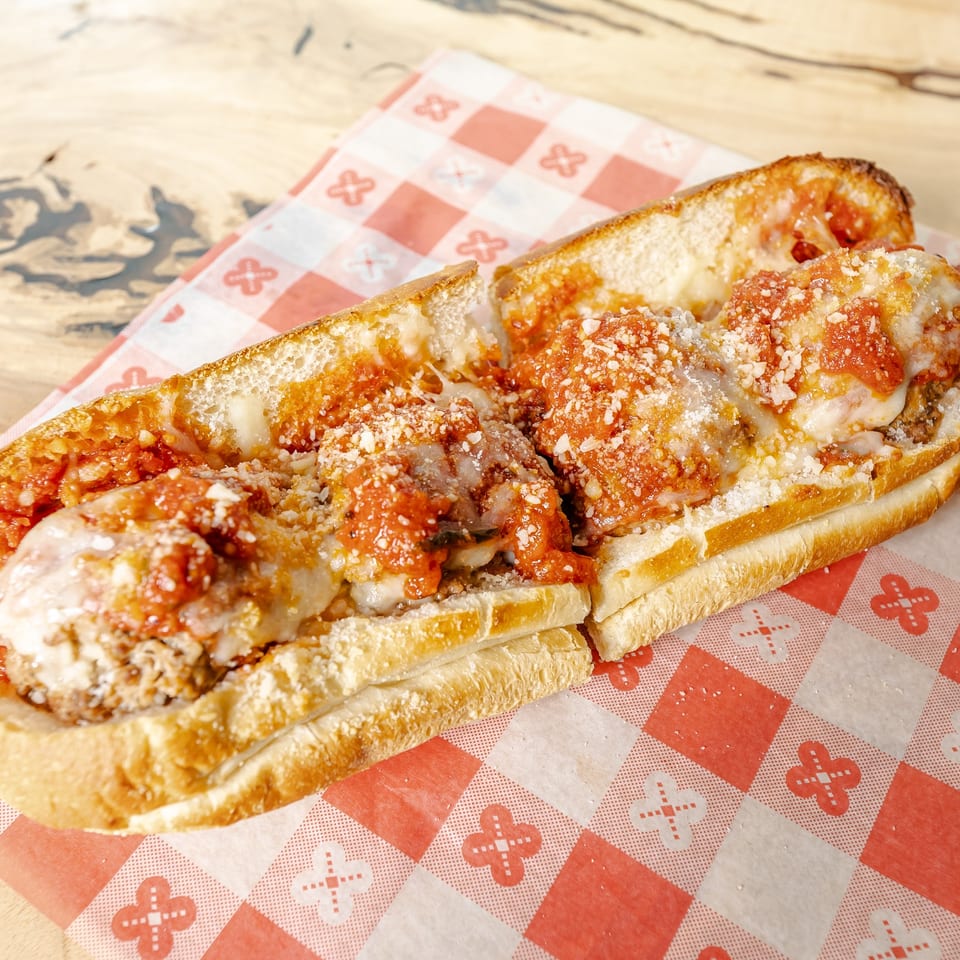 Toasted meatball sub topped with tomato sauce, melted cheese, and grated Parmesan on red-and-white checkered paper.