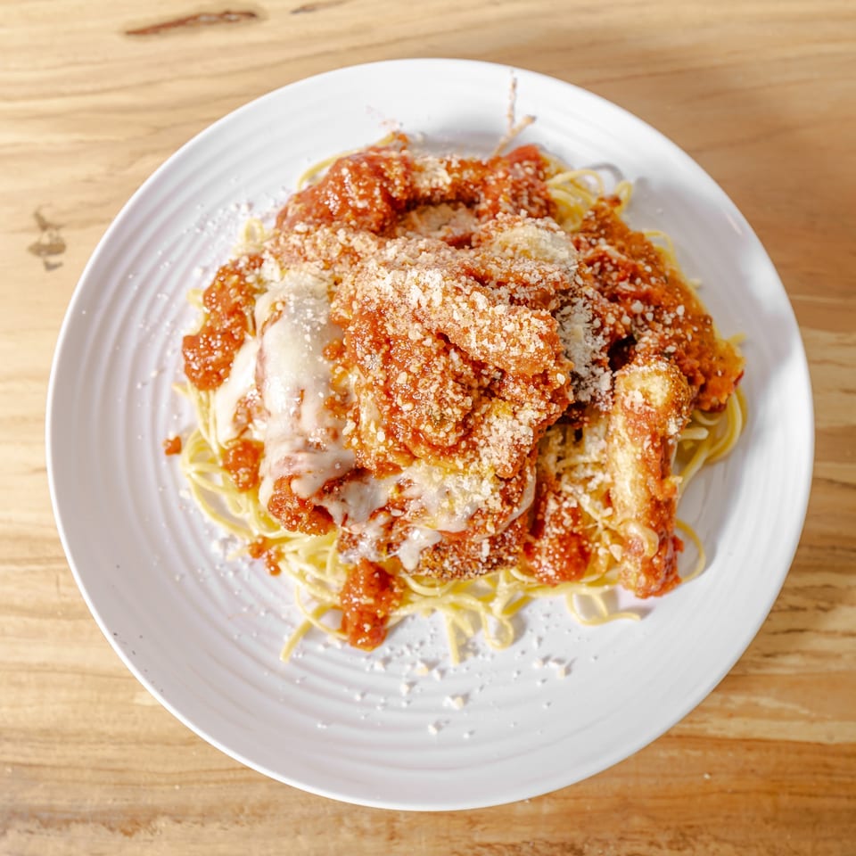 Spaghetti topped with breaded chicken, marinara sauce, melted cheese, and grated Parmesan on a white plate.