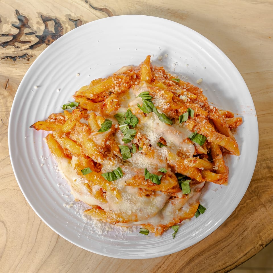 Baked penne pasta in tomato sauce topped with melted cheese, grated Parmesan, and fresh basil on a white plate.