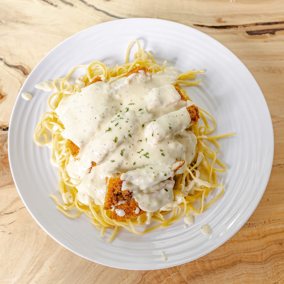 Breaded chicken topped with creamy white sauce and herbs, served over spaghetti on a white plate.