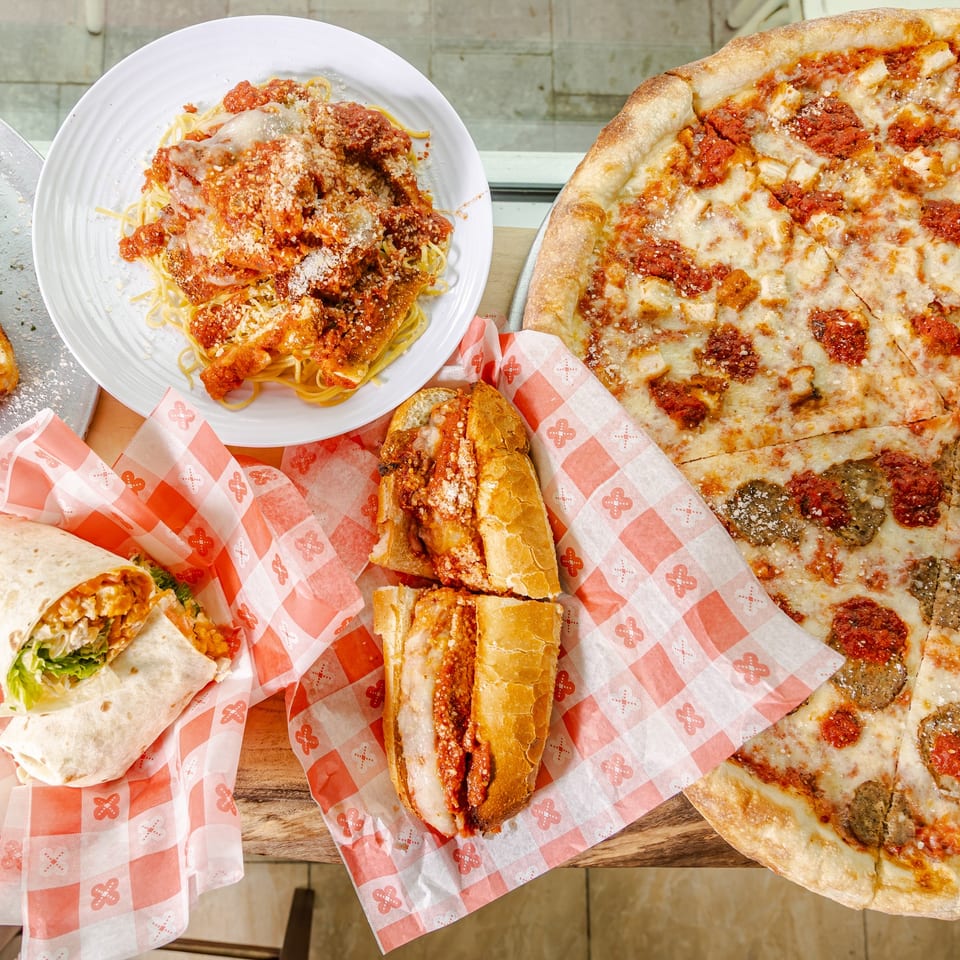 Overhead spread of cheesy pizzas, spaghetti with meatballs, chicken parmigiana sandwiches, and a chicken wrap on checkered paper.