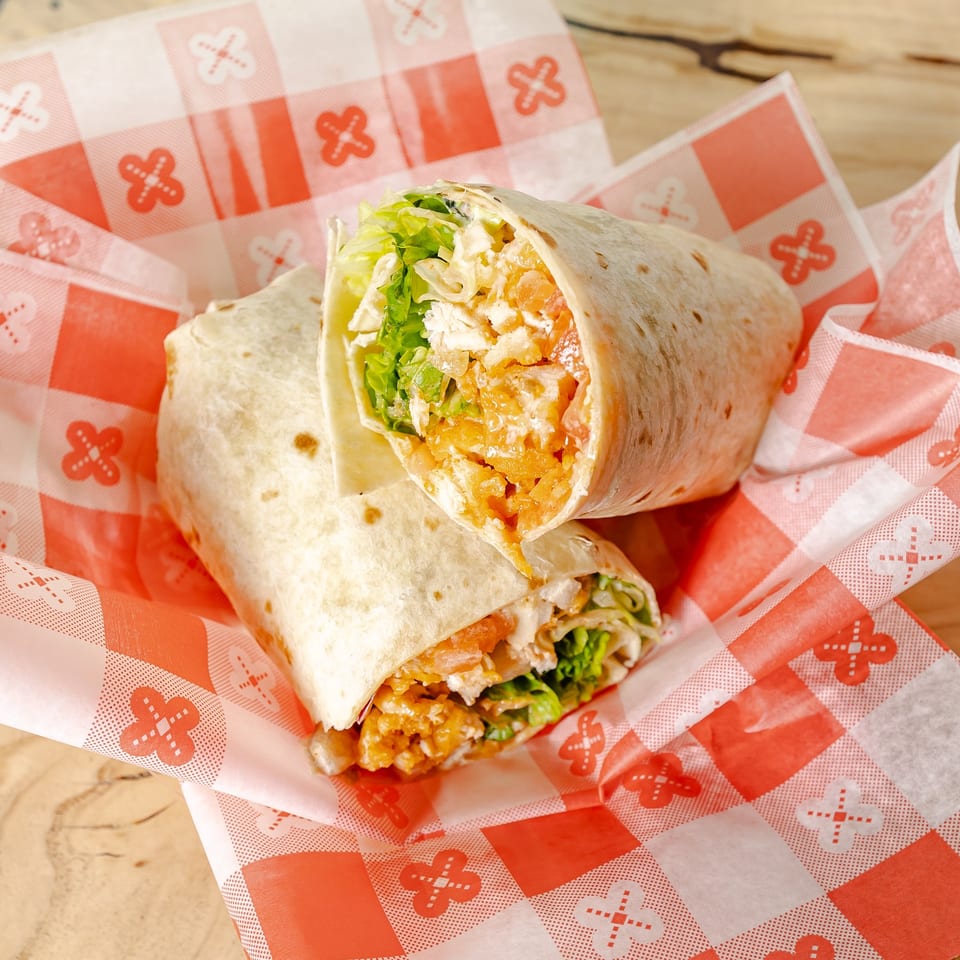 Two toasted chicken wraps filled with lettuce, crispy coating, cheese, and sauce, served on patterned paper over a wooden table.