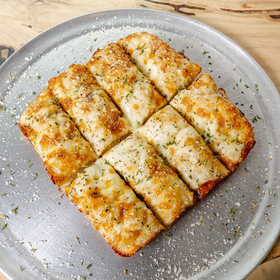 Golden-brown cheesy breadsticks sprinkled with herbs and grated cheese, arranged on a metal platter atop a wooden table.